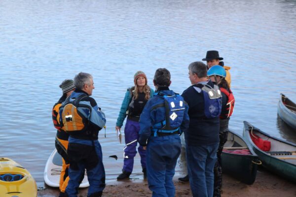 A female coach talks to a group of six paddlers at the bank of the river. Coach Award eLearning