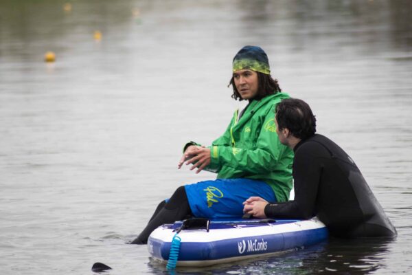 A coach sits on a standup paddle board whilst explaining something to another coach. Coach Developer elearning.