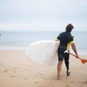 A paddle on a beach, walking towards the sea with a standup paddle board under their left arm. Introduction to Disability Awareness